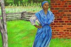 African-American woman in long dress and head scarf in front of a brick building and rural field.