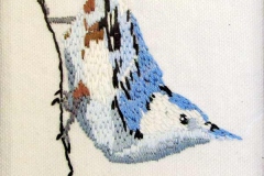 Stitched blue and white bird perched diagonally on a thin branch.