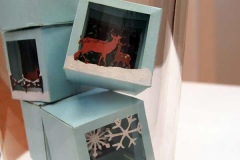 Detail up close: pale blue colored ice cubes, each containing a snow scene, snow flakes, rein deer, wooden fence.