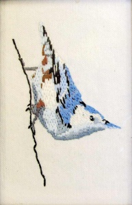 Stitched blue and white bird perched diagonally on a thin branch.