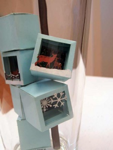 Detail up close: pale blue colored ice cubes, each containing a snow scene, snow flakes, rein deer, wooden fence.