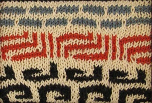 Beige knitted art with back swirls and red and gray geometric shapes.