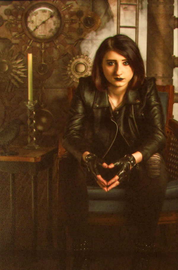 Young, brown haired woman in leather jacket and driving gloves sitting in front of an antique clock.