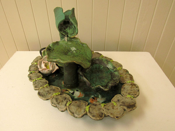 Green ceramic fountian with running water splashing over coy fish amid lilly pad edged basin.